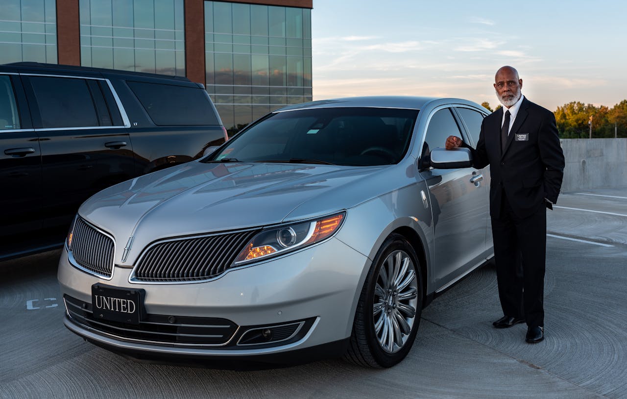 about-us Professional chauffeur in a suit standing next to a luxury car in outdoor setting.